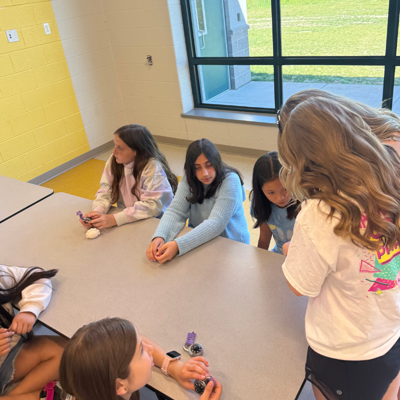 Several young girls sit around a table, looking at small objects in their hands.