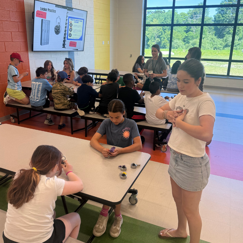 Students practice opening lockers in a school commons area, with instructions displayed on a large screen.