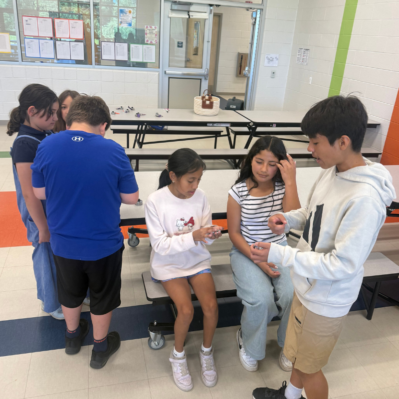A group of students gather around a table in a school cafeteria, looking at small objects.