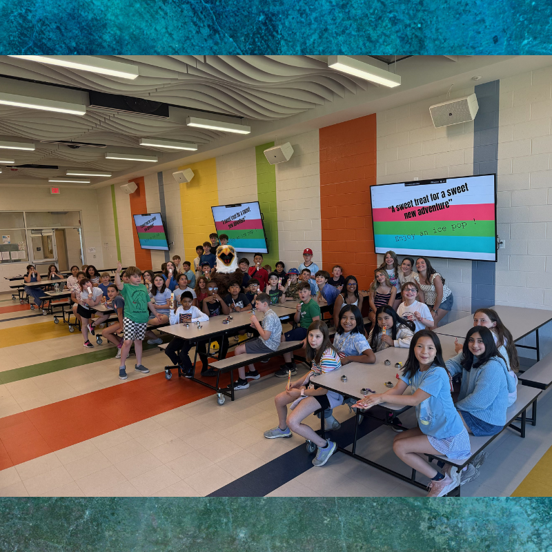 A group of students sits at tables in a colorful cafeteria, with a large screen displaying "A sweet treat for a sweet new adventure" and "Enjoy an ice pop!".