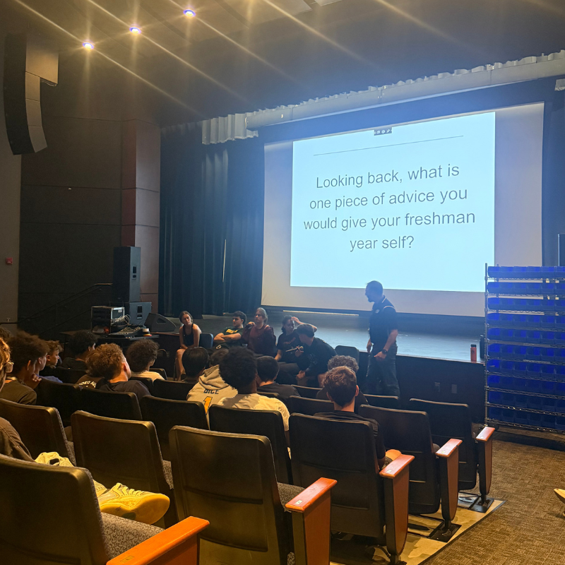 A group of students sits in an auditorium facing a large screen with the text 'Looking back, what is one piece of advice you would give your freshman year self?'.