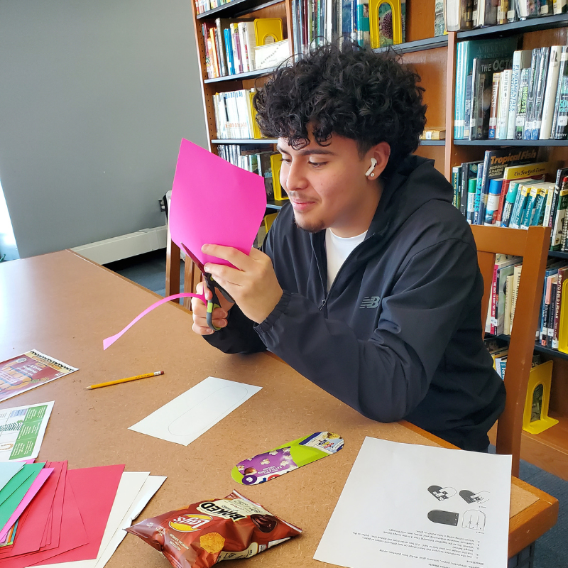 A young person with curly hair cuts pink paper with scissors at a table in a library.