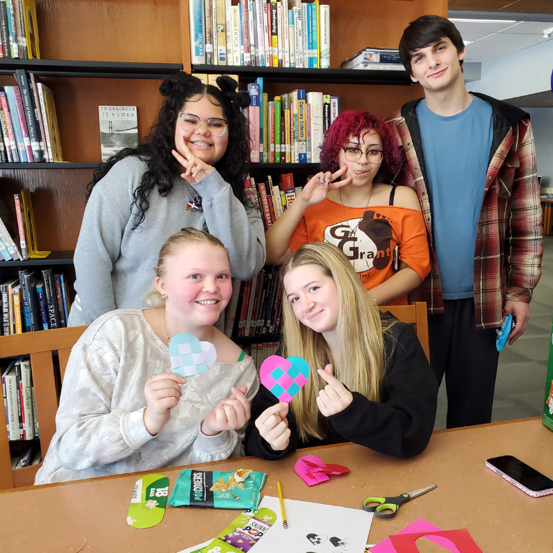 Five young people pose for a photo in front of bookshelves, holding handmade paper hearts.