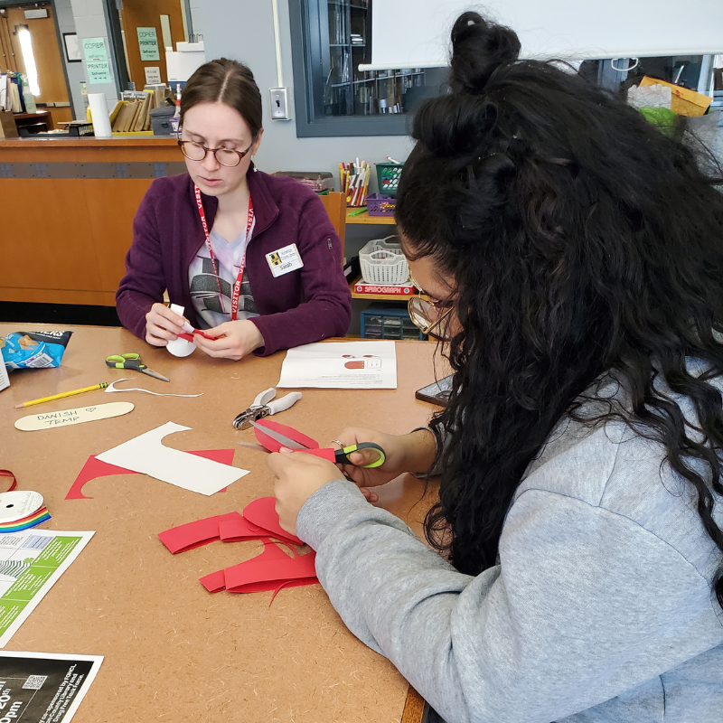 Two people are crafting at a table, cutting red paper with scissors.
