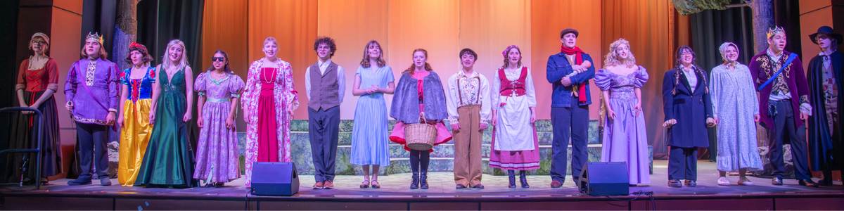 A diverse cast of performers in colorful costumes stand on a stage, posing for a photo.