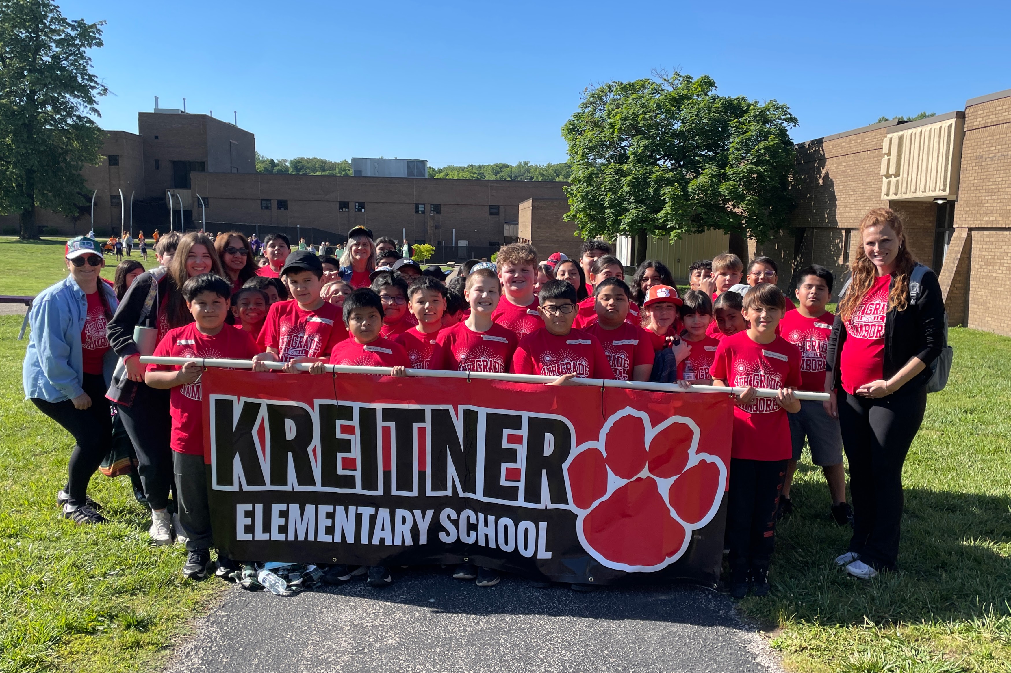 A group of students and adults in red shirts stand together holding a banner that reads 'KREITNER ELEMENTARY SCHOOL'.