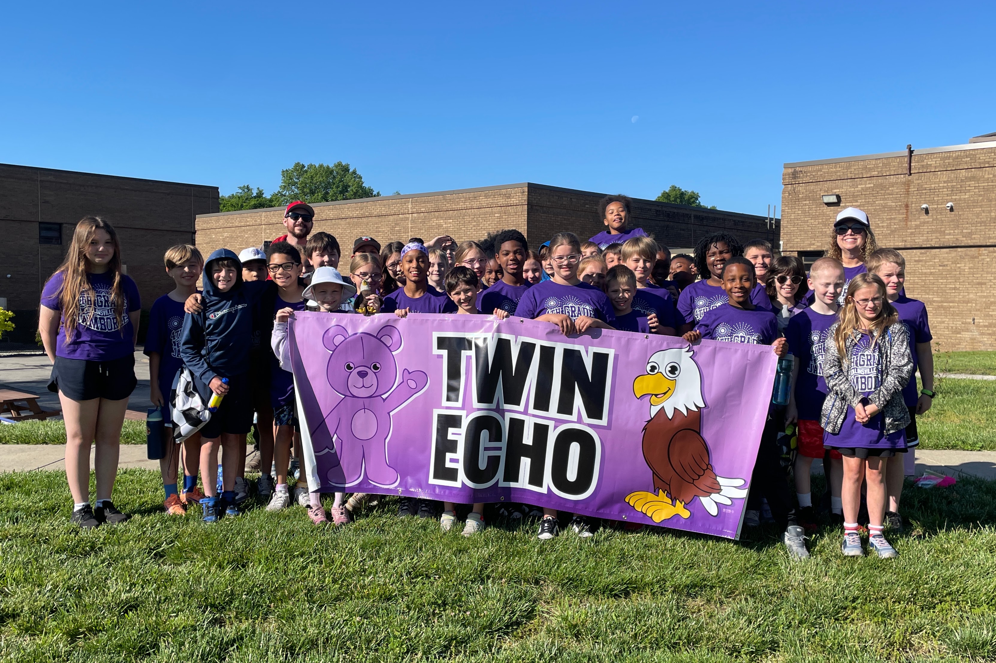 A large group of children and adults stand together outdoors, holding a purple banner that says "TWIN ECHO" with cartoon characters.