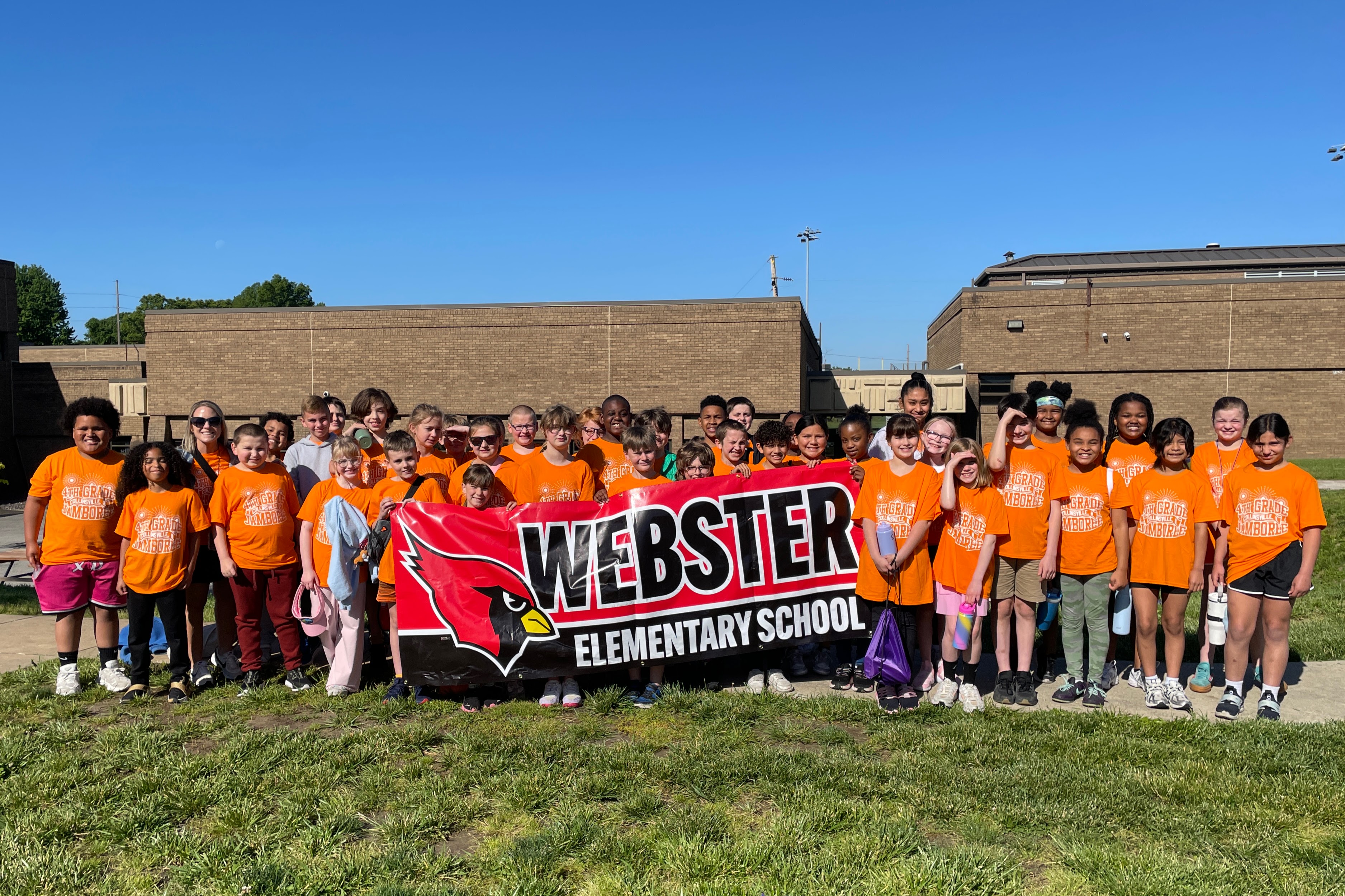 A large group of students and adults in orange shirts stand together outdoors, holding a banner that reads 'WEBSTER ELEMENTARY SCHOOL'.