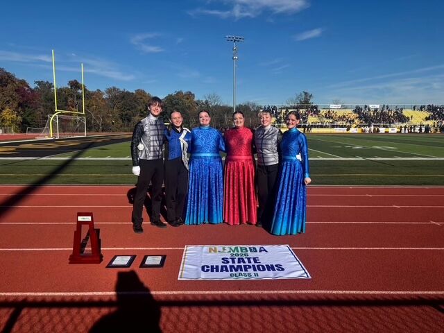 Six band members in uniforms stand proudly on a track, with a 'State Champions' banner and trophies in front of them on a sunny day.