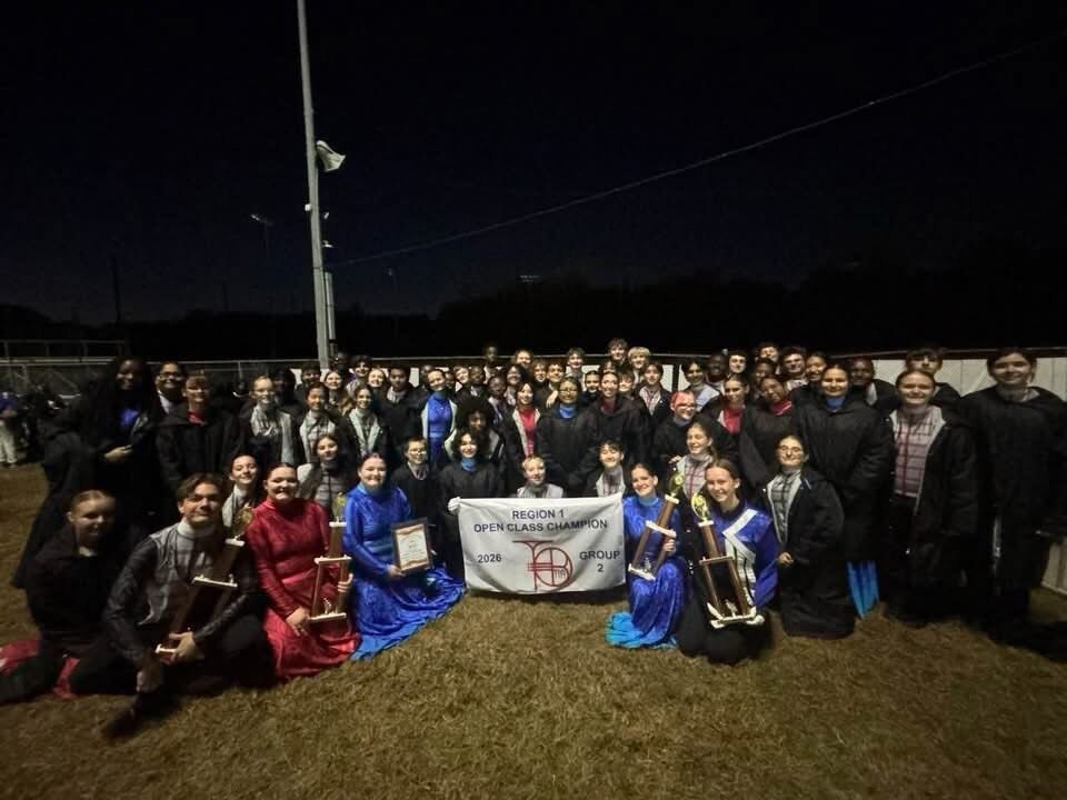 A large group of marching band members poses outdoors at night, holding trophies and a banner that reads 'Region 1 Open Class Champion 2026'.
