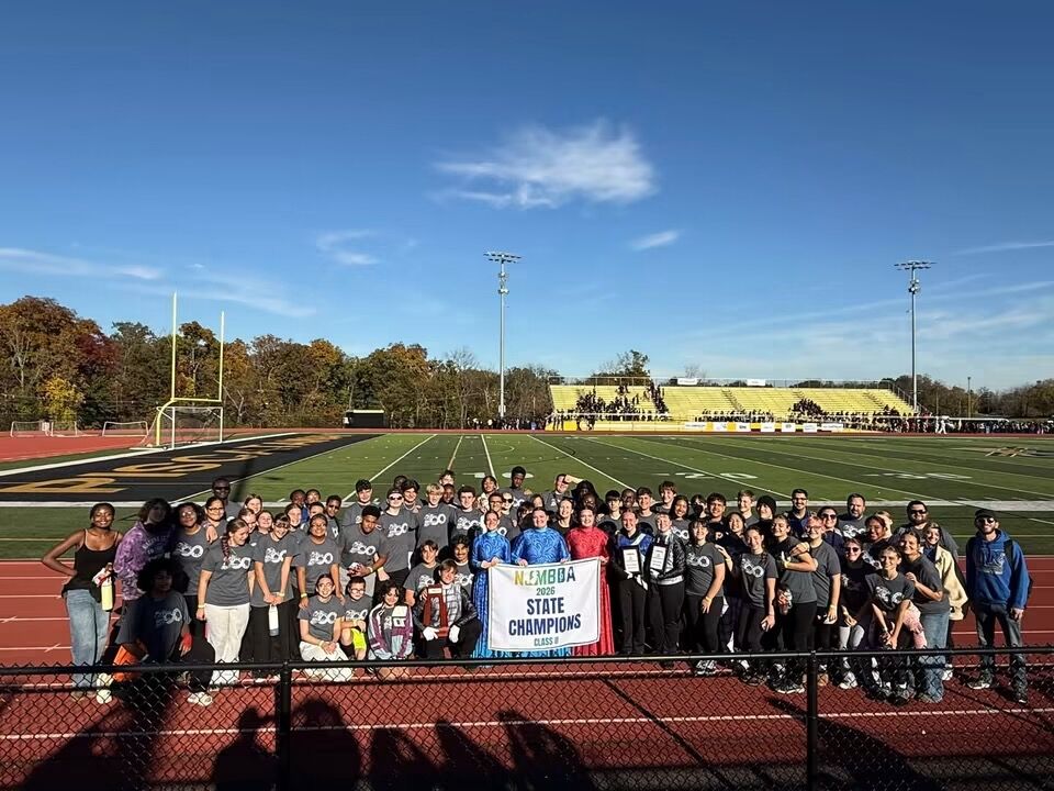 A large group of students in grey shirts stand proudly on a track, holding a "STATE CHAMPIONS" banner on a sunny day.