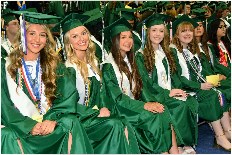 A group of smiling graduates in green gowns and caps sit together at a ceremony.