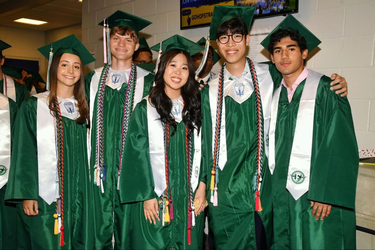 Five graduates in green gowns and caps smile for a photo, adorned with colorful cords.