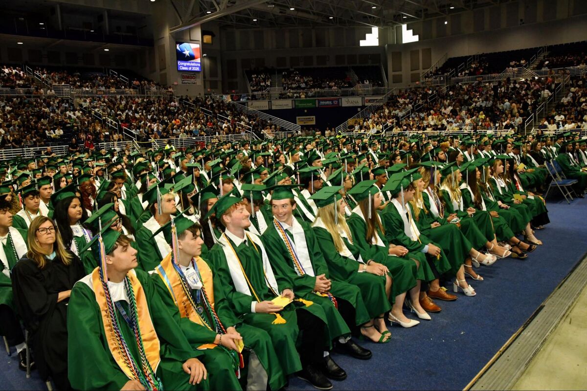 Graduates in green gowns and caps sit in an arena, facing forward for a ceremony.