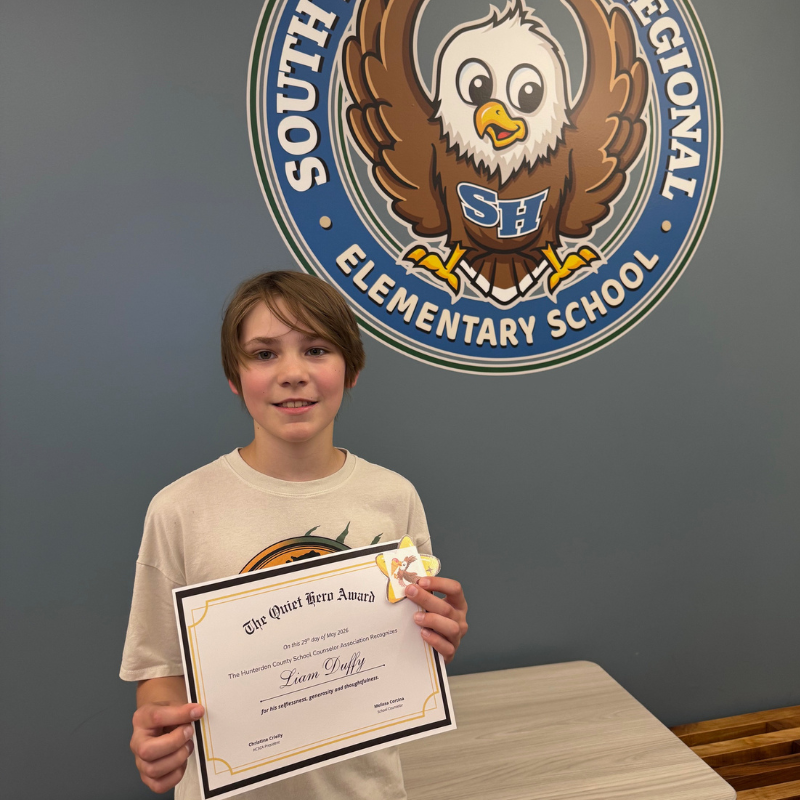 A smiling boy holds a certificate for the "Quiet Hero Award" in front of a school logo.