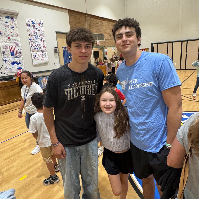 Three young people pose for a photo indoors, with two older boys flanking a smiling girl.