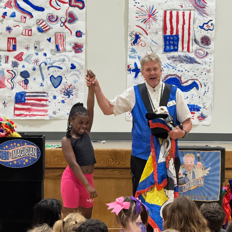 A magician holds a rabbit from a hat while a young girl smiles on stage.