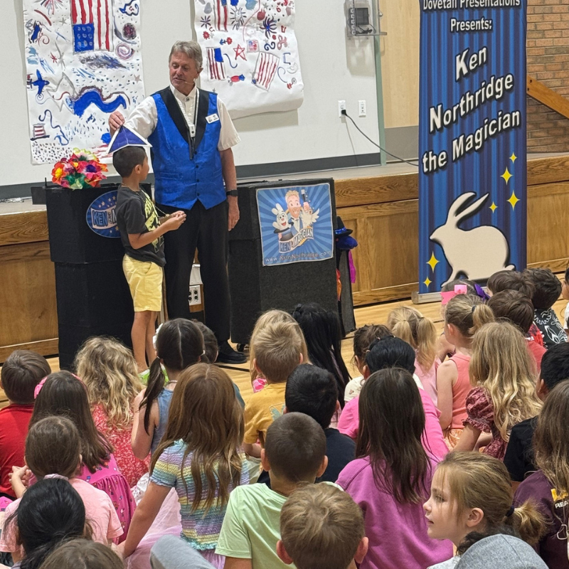 A magician in a blue vest interacts with a young boy on stage in front of an audience of children.