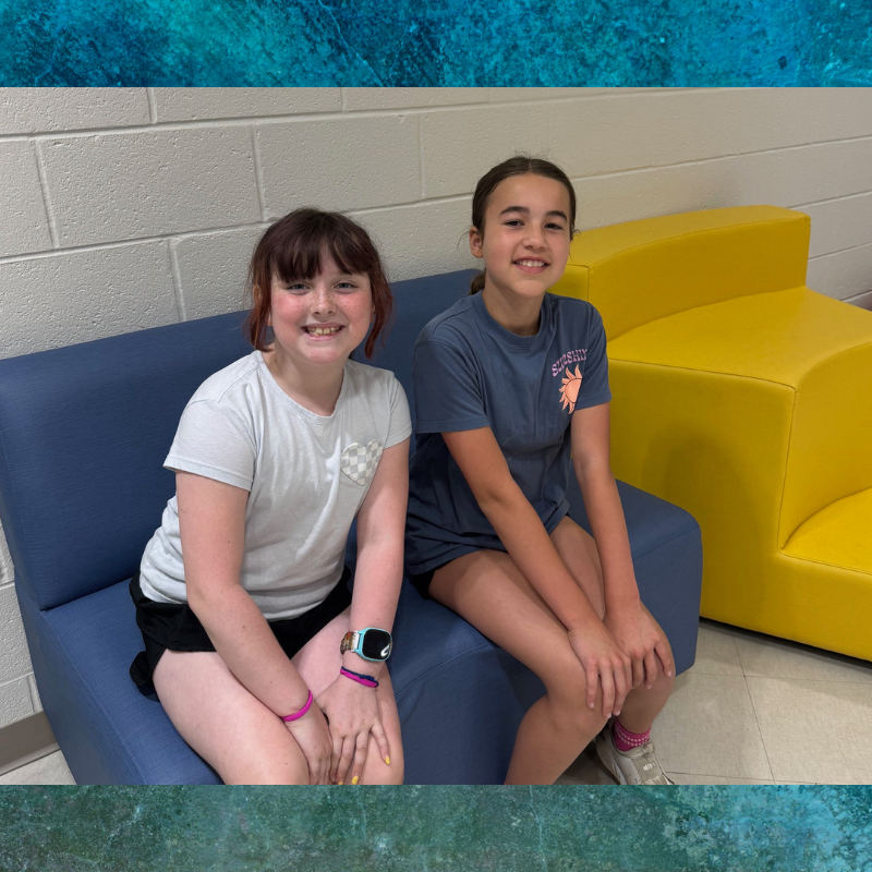 Two smiling young girls sit side-by-side on a blue couch in front of a brick wall.