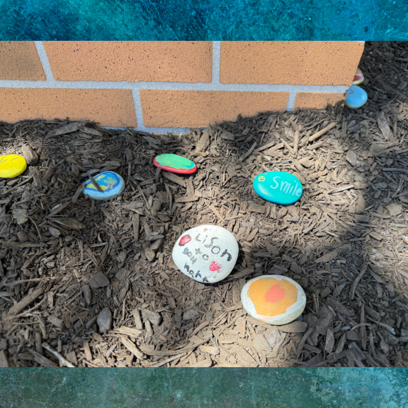 Painted rocks with positive messages are scattered on mulch near a brick wall.