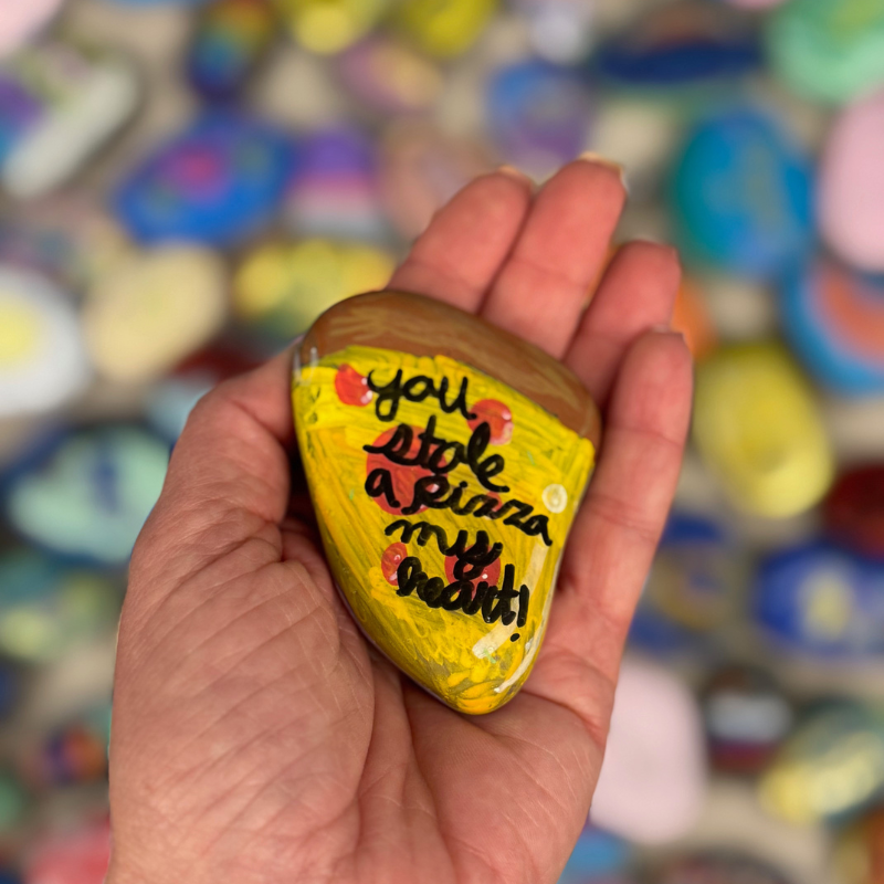 A hand holds a painted rock shaped like a pizza slice with the text 'you stole a pizza my heart!'.