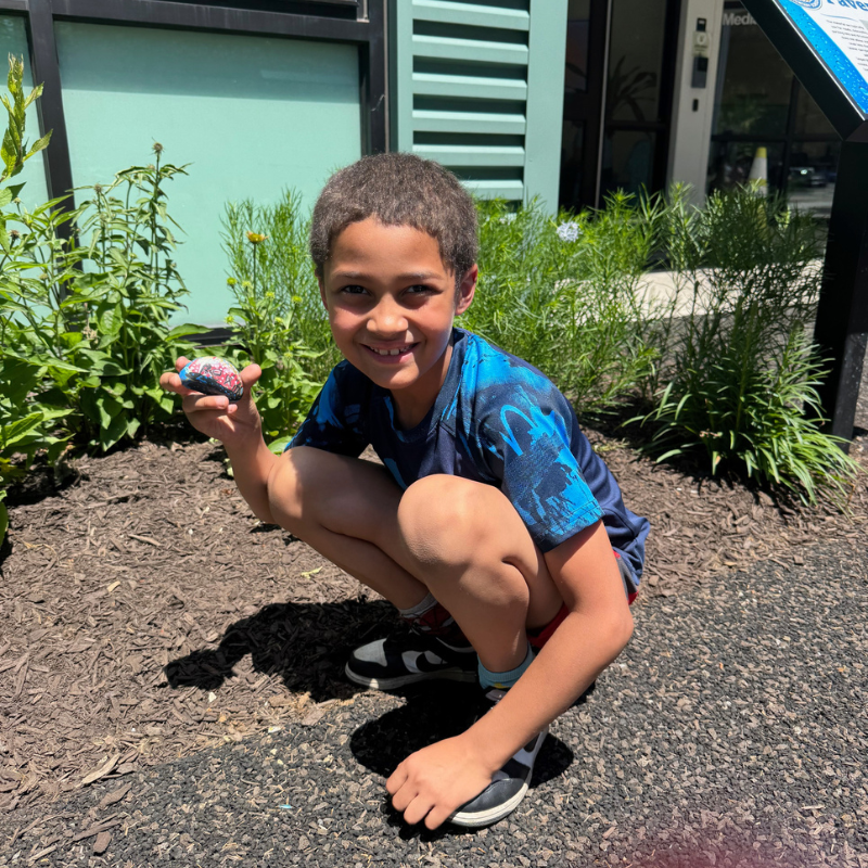 A young boy smiles while holding a painted rock in a garden area.