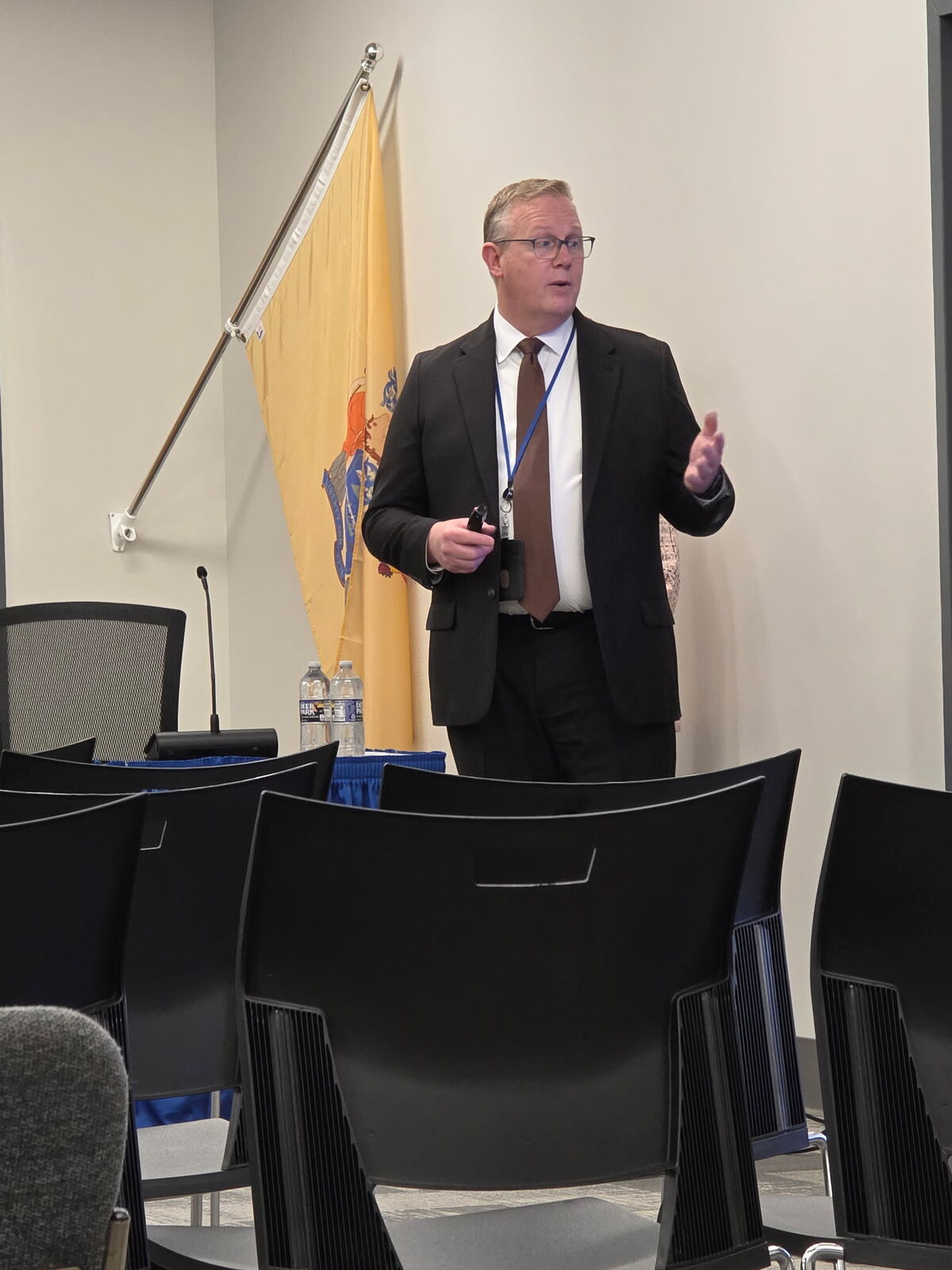 A man in a suit and tie gestures while speaking in front of a flag and chairs.