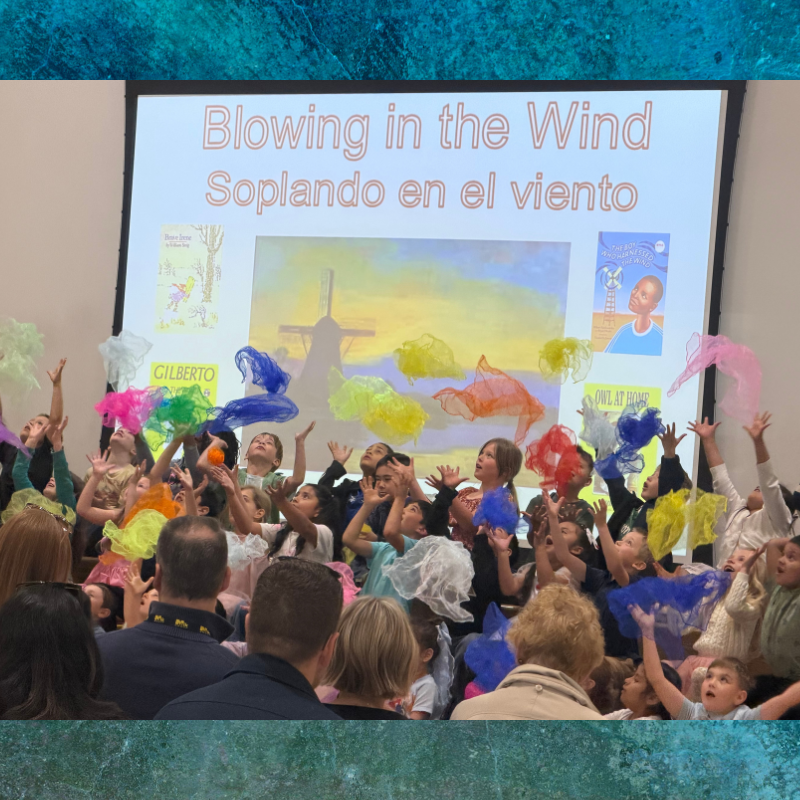 Children joyfully toss colorful scarves in the air during a performance with "Blowing in the Wind" displayed on a screen.