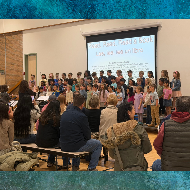 A group of children stand on risers, singing in front of a projector screen displaying "Read, Read, Read a Book" and a list of favorite books.