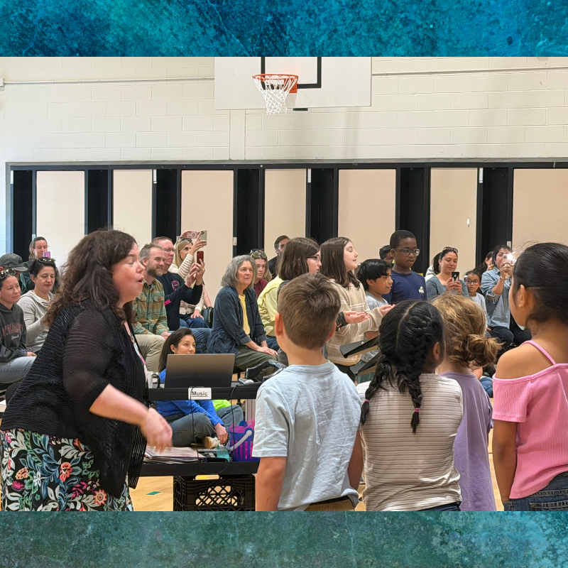 A group of children and adults are gathered in a gymnasium, with a woman in the foreground appearing to lead them.