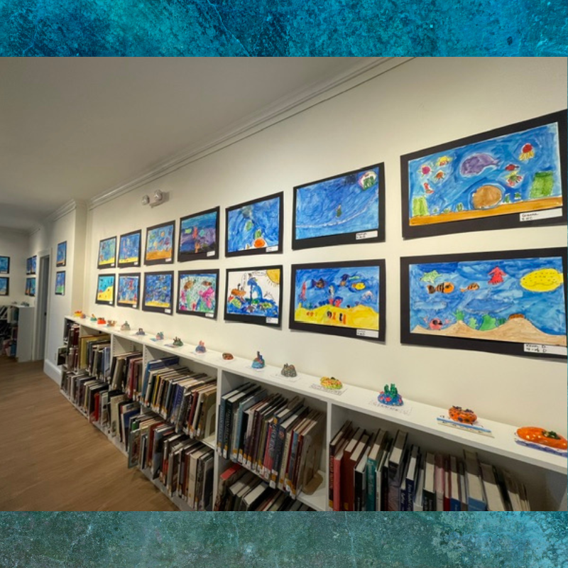 A wall displays rows of colorful children's artwork depicting underwater scenes, above bookshelves filled with books.