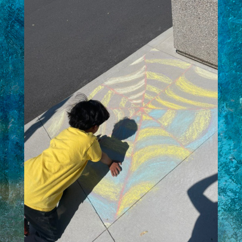 A child in a yellow shirt draws a colorful spiderweb with chalk on a sidewalk.