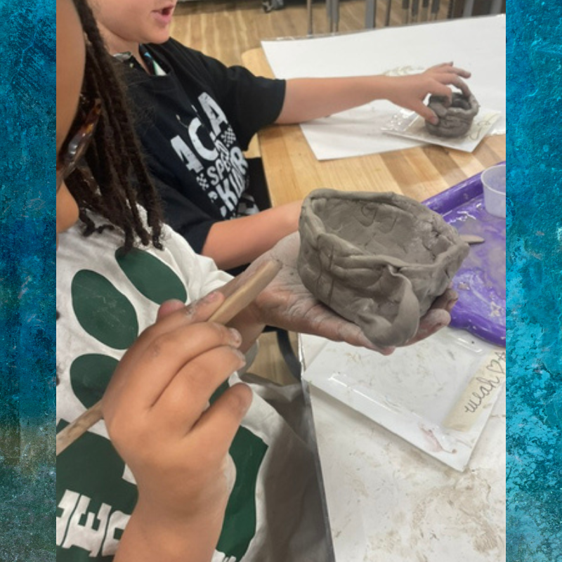 A child's hands shape a clay bowl with a wooden tool in an art class.