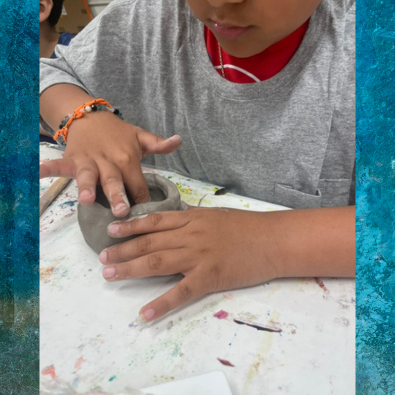 A child's hands shape a small clay pot on a white table.