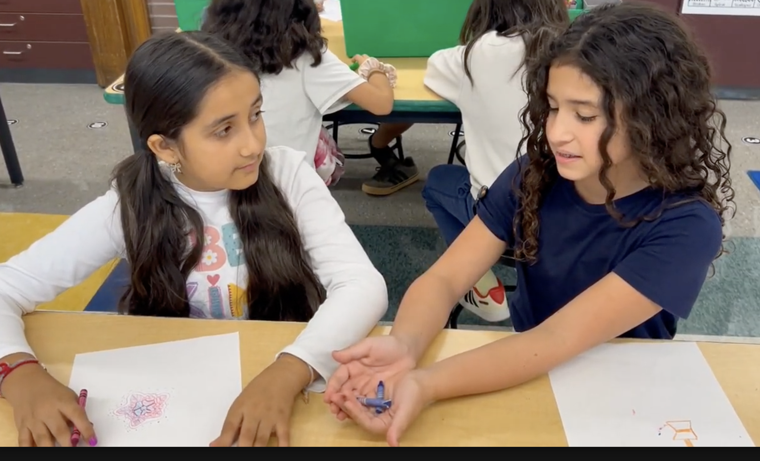 Two young girls sit at a classroom desk, sharing blue crayons while working on their drawings.
