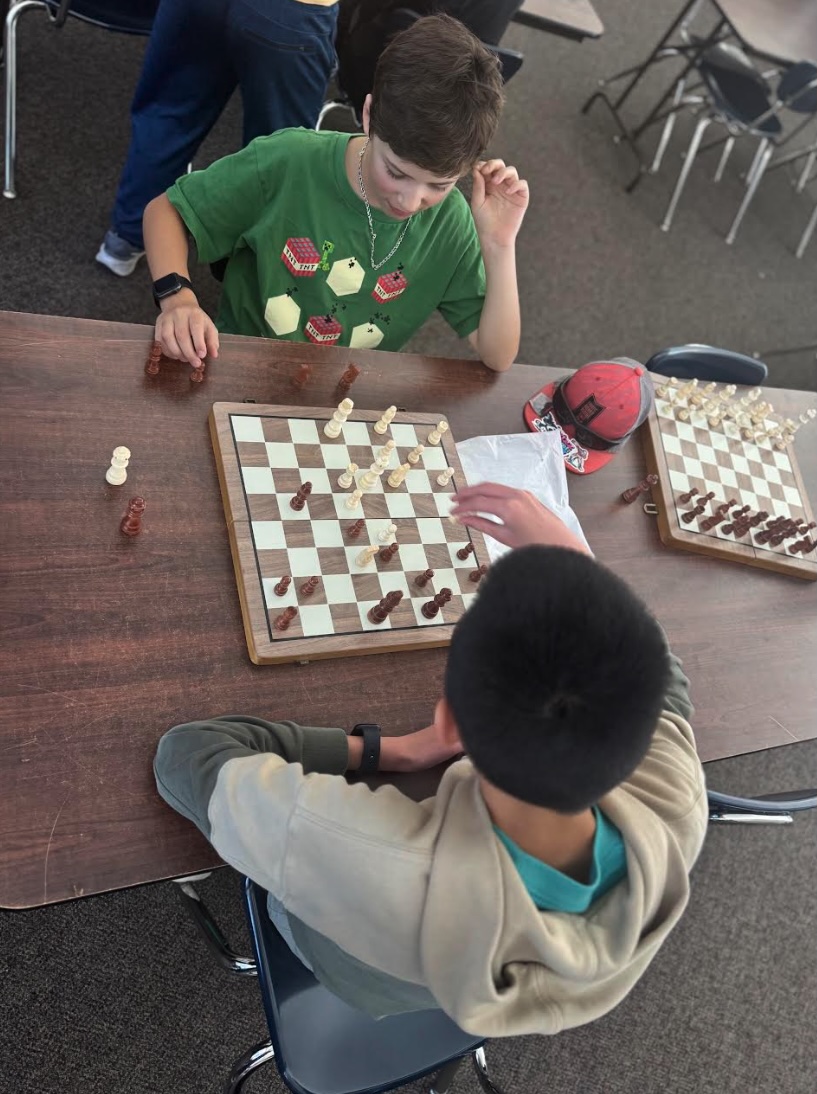 Two boys concentrate on a chess game at a wooden table, with chessboards and pieces visible.