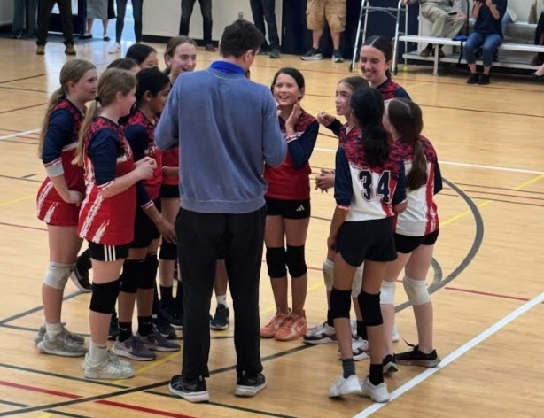 A group of RMS female volleyball players in red and blue uniforms huddle around their coach on a wooden court.