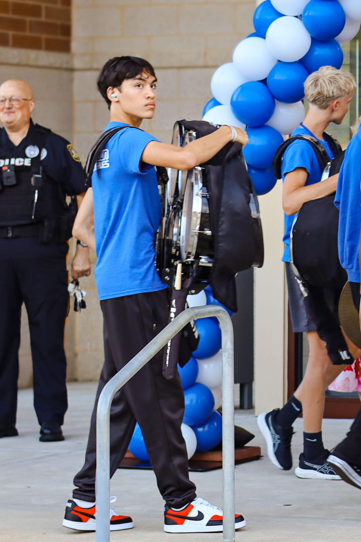 A young drummer in a blue shirt carries a drum set outside, past a police officer and blue-and-white balloons.