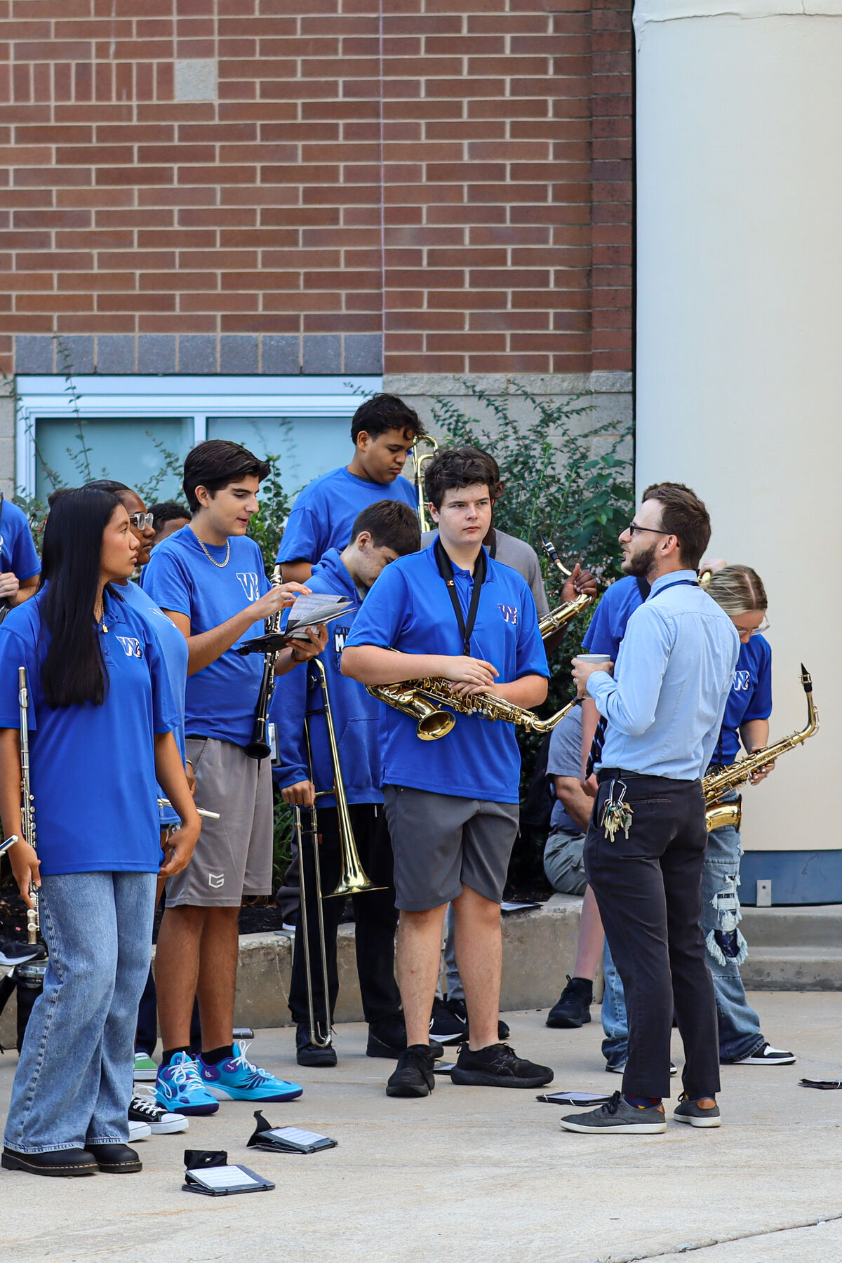 A diverse high school marching band in blue shirts prepares to practice outside a brick building.