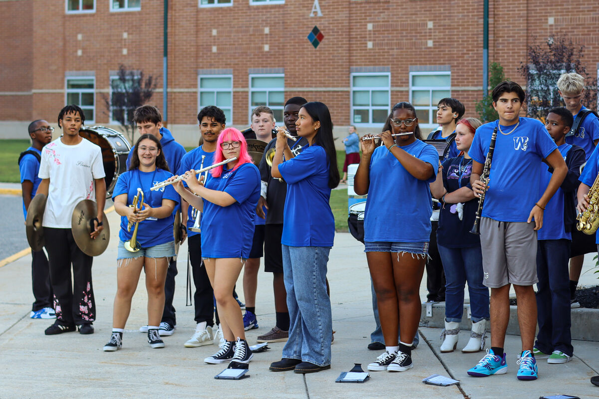 A diverse high school marching band in matching blue shirts and white t-shirts stands outside a brick school building.