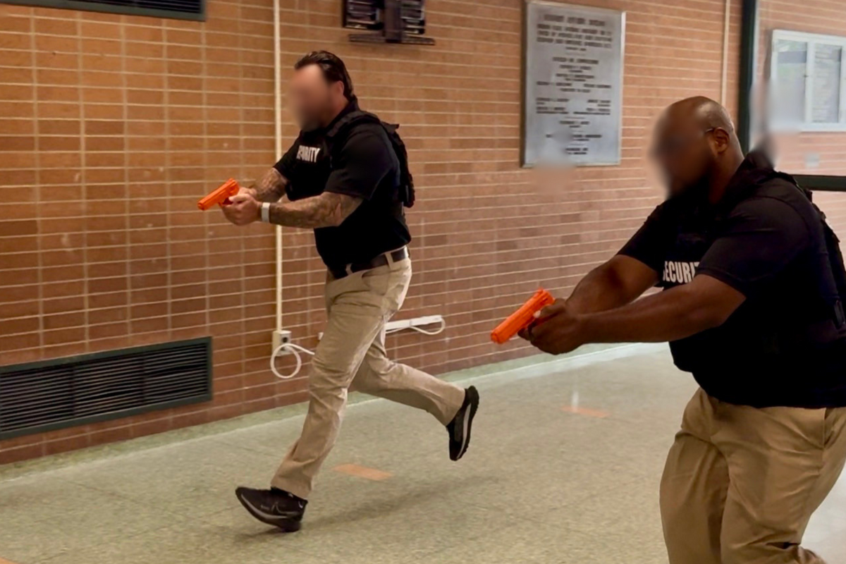 Two security personnel in tactical vests practice with orange training firearms in a hallway.