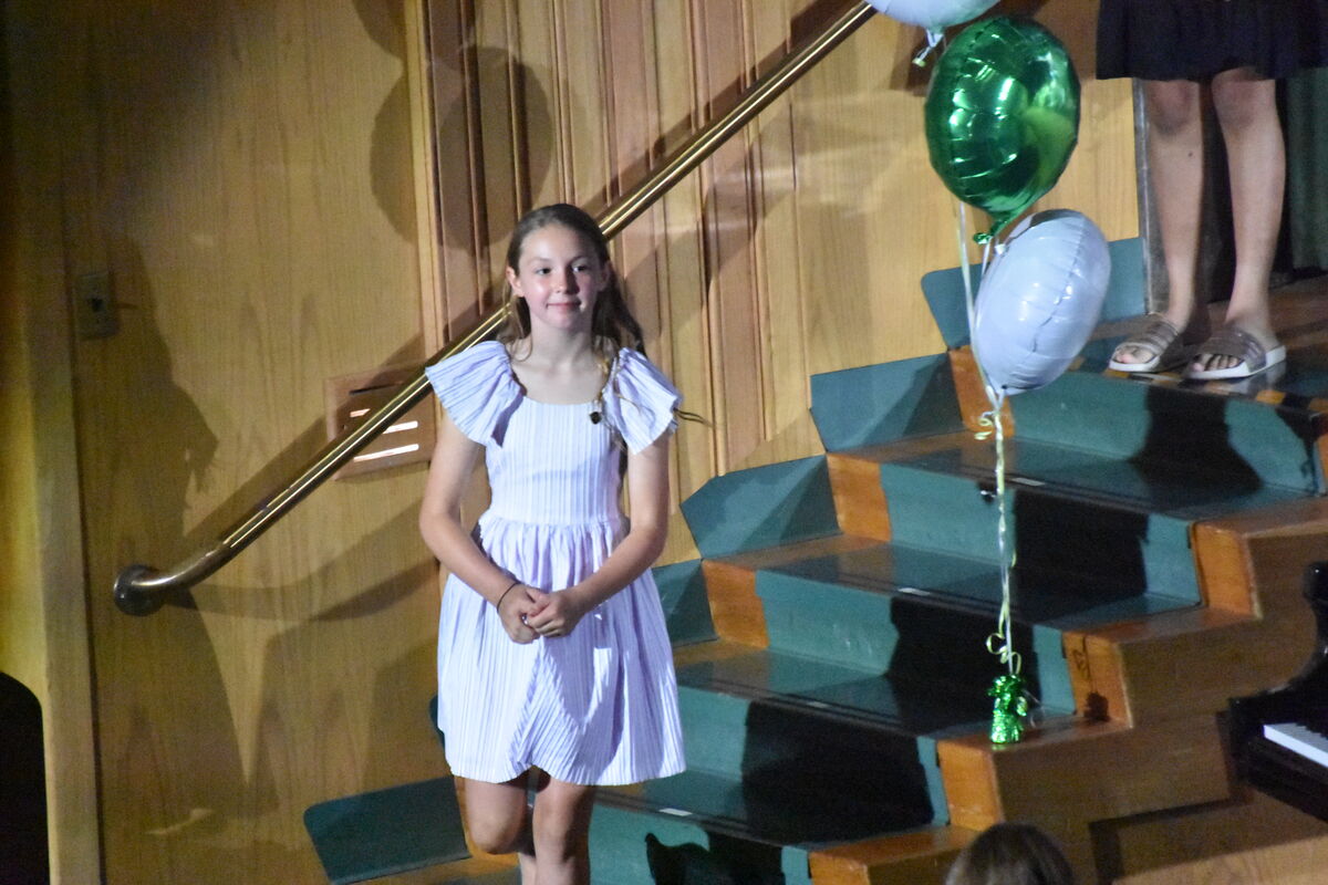 A young girl in a light purple dress stands on a stage with a wooden wall and a railing.
