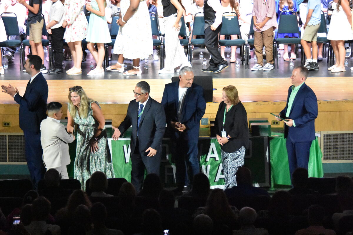 Adults in suits and dresses stand on a stage, with a group of children in formal attire behind them.