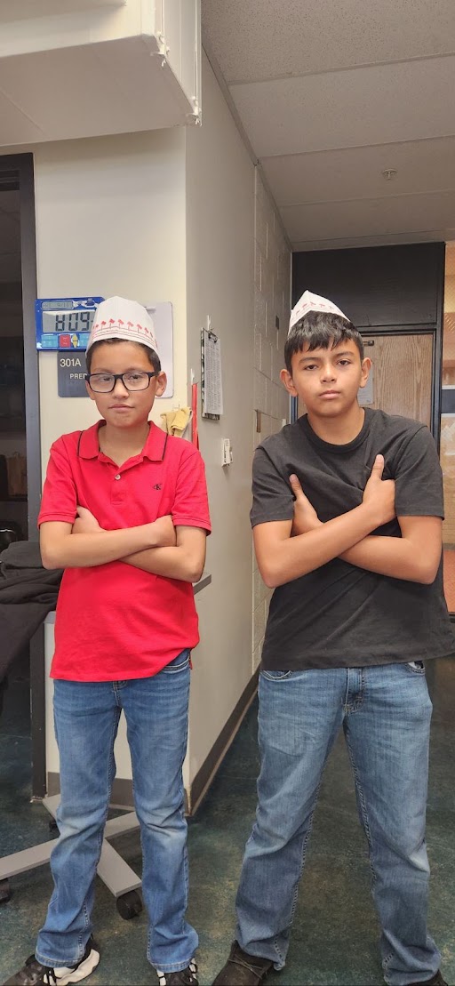 Two boys wearing paper hats stand in a hallway with their arms crossed, posing for the camera.