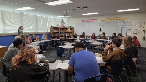 A teacher stands before a group of students seated in a large circle of desks inside a bright classroom.
