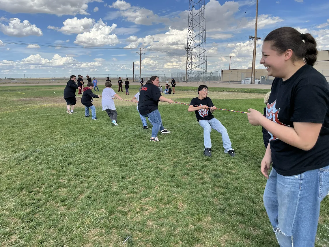 A group of students enthusiastically participate in a game of tug-of-war on a grassy field under a cloudy blue sky.