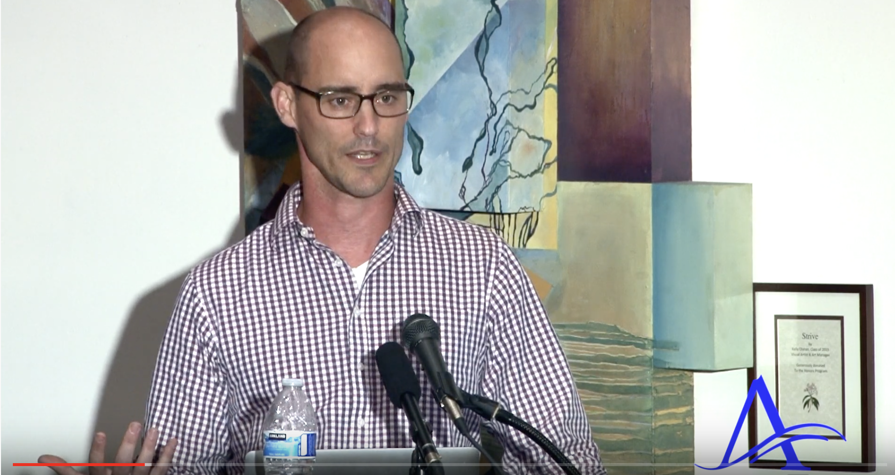Photograph of Kevin Timpe talking in front of abstract art. He's wearing a lovely plaid shirt. Bottle water and two microphones are visible in front of him.