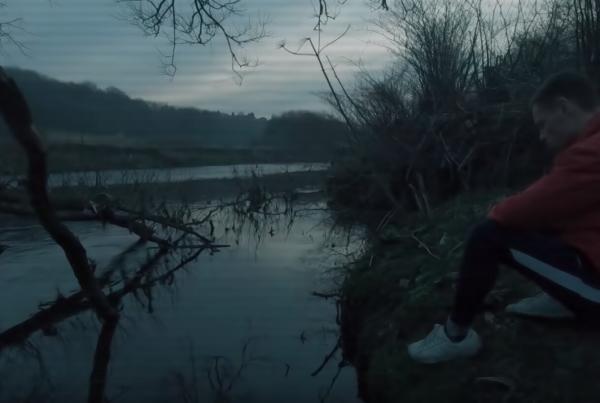 a still from the video, featuring a man in a red hoody sitting on the ground by a lake