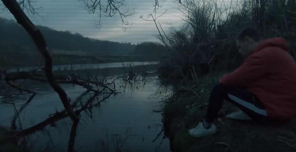 a still from the video, featuring a man in a red hoody sitting on the ground by a lake