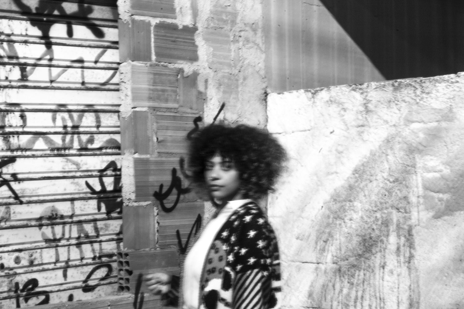 Fotografia em preto e branco da artista Thati Dias. Ela é uma mulher negra, de cabelos crespos encaracolados e veste blusa estampada.